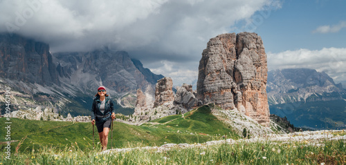 Smiling female trekker walking with backpack and trekking poles by green mountain hill with picturesque Dolomite Alps Cinque Torri formation on the background. Active people and mountains concept.