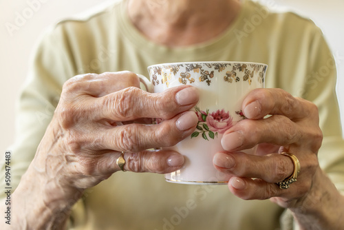 Image of an elderly woman with arthritis in her hands trying to hold a beautiful tea cup. Her fingers are twisted and contrast against the delicate tea cup.