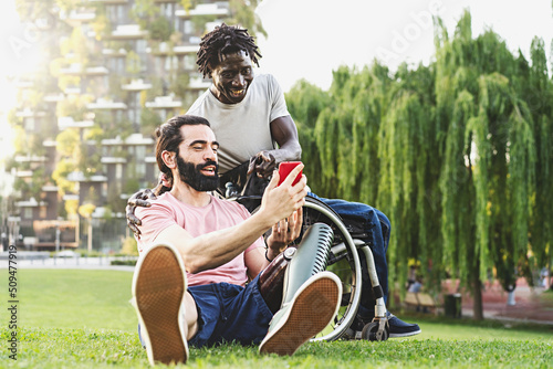Two friends, one Hispanic with a prosthetic leg and one African in a wheelchair are relaxing outdoors chatting together and using smartphones and social networks - disability and integration concept