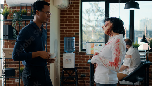 Hungry creepy zombie talking with businessman while dead coworker covered in blood walks by. Person discussing with bizarre looking evil undead monster in office while mindless colleague passes by.