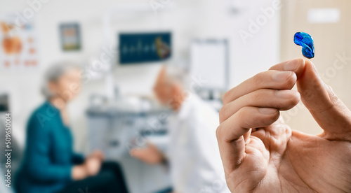Intra-ear hearing aid in doctor hands, close-up. Selection of hearing aids for senior female patient with audiologist over background, soft focus