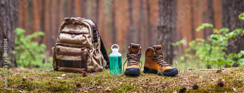 Hiking and camping equipment in forest. Backpack, water bottle and leather ankle boots. Panoramic view with copy space 