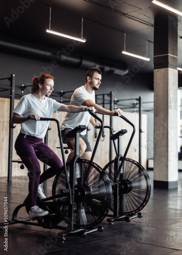 Athletic man and woman doing intense workout together by pedaling air bike in modern gym. Healthy lifestyle
