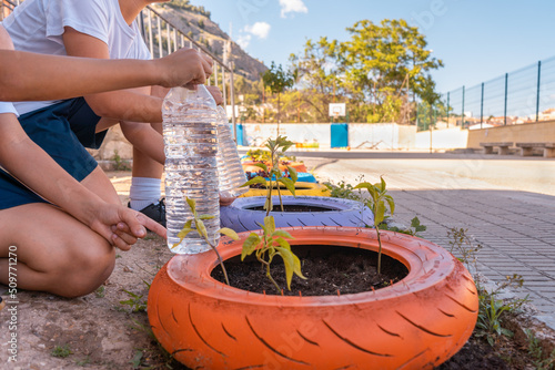 Dos niños irreconocibles regando las plantas con botellas de agua en el patio del colegio. Fotografía horizontal con espacio para texto.