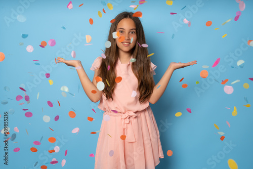 Portrait of a happy beautiful girl standing under confetti rain and celebrating isolated over blue background