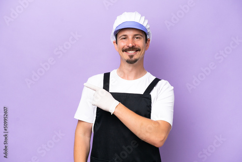 Fishmonger man wearing an apron isolated on purple background pointing to the side to present a product