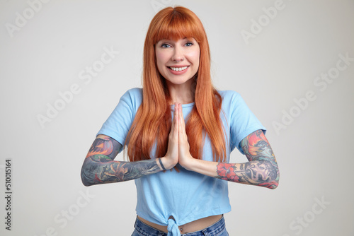 Indoor portrait of young ginger female posing over white wall pressed he palm in praying position with broad smile on her face