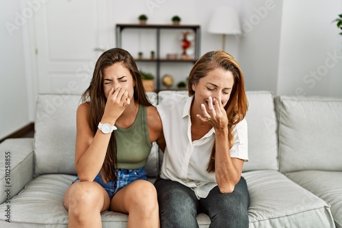 Mother and daughter together sitting on the sofa at home smelling something stinky and disgusting, intolerable smell, holding breath with fingers on nose. bad smell