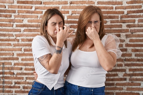 Hispanic mother and daughter wearing casual white t shirt smelling something stinky and disgusting, intolerable smell, holding breath with fingers on nose. bad smell