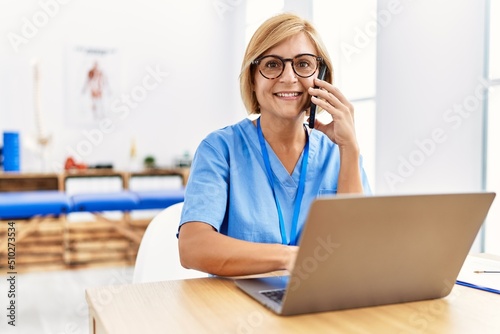Middle age blonde woman wearing physio therapy uniform talking on the smartphone and using laptop at clinic