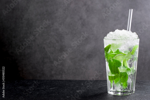 Cold mojito with lime and mint leaves on black table. Fresh beverage with crushed ice on top and straw in glass made of citrus fruit c with copy space