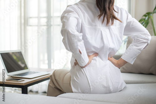 Working woman sitting on the sofa in the living room at home She used both hands to press down on the lower back. She is suffering from back pain from sitting for a long time.