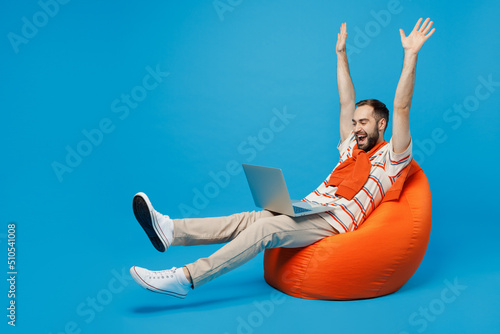Full body young excited fun cool man 20s in orange striped t-shirt sit in bag chair hold use work on laptop pc computer with overstretched hands legs isolated on plain blue background studio portrait
