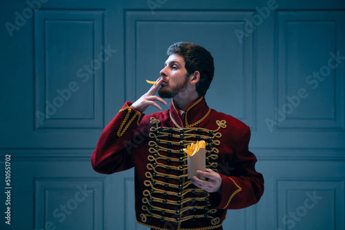 Comic portrait of young man in image of medieval hussar smoking fries isolated on dark blue background. Retro style, comparison of eras concept.