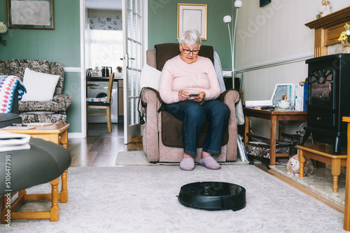 Senior woman using a robot vacuum cleaner through phone settings while sitting on armchair at home. Mature older lady holding mobile phone and control cleaning. Smart home. Seniors and technology.