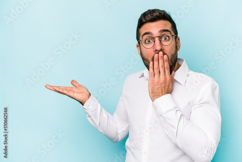 Young caucasian man isolated on blue background impressed holding copy space on palm.