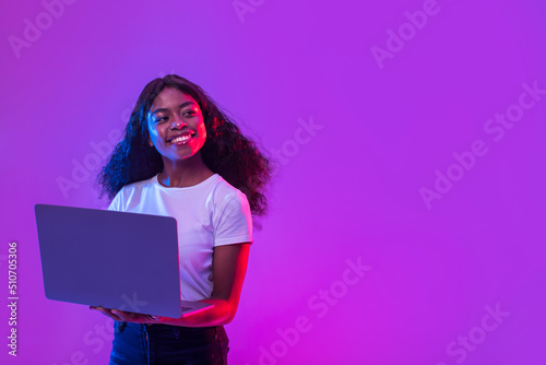 African American lady using laptop for online work, education or communication, looking at empty space in neon light
