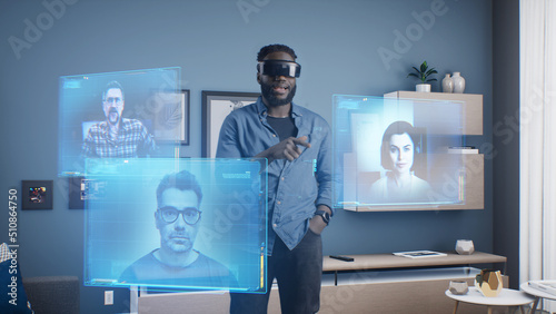 Afro-american man wearing VR goggles at online business meeting in meta universe cyberspace talking with colleagues over video conference, holographic windows in front of him