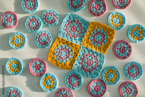 Top view of multicolored crochet motifs - one big square and many small circles on white surface with rough texture, bold shadows in hard light.