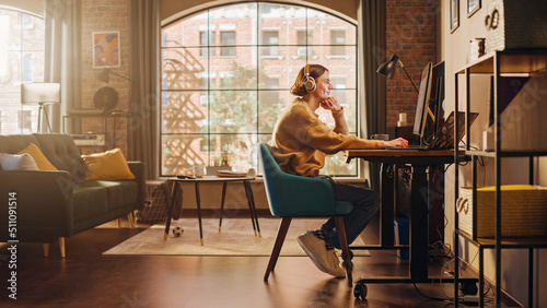 Young Handsome Man Working from Home on Desktop Computer in Sunny Stylish Loft Apartment. Creative Designer Wearing Cozy Yellow Sweater and Headphones. Urban City View from Big Window.