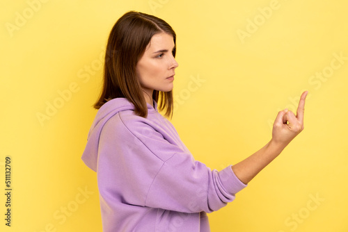 Side view portrait of rude vulgar woman showing middle fingers, impolite gesture of disrespect, looking with aggression hate, wearing purple hoodie. Indoor studio shot isolated on yellow background.