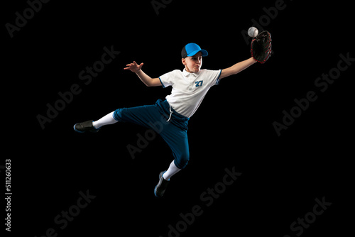 Dynamic portrait of little baseball player, pitcher in blue-white uniform training isolated on black studio background. Concept of sport, achievements, studying, competition