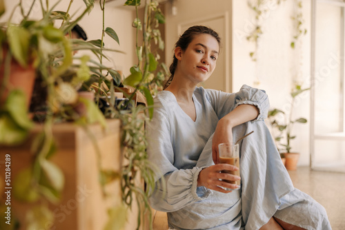 Pretty young caucasian woman with coffee smoothie enjoys pastime sitting indoors. Brunette wears blue sundress on summer day. Relaxation concept