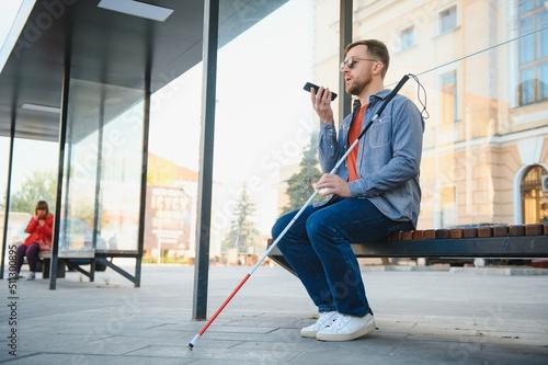 blind man with white cane waiting for public transport in city.