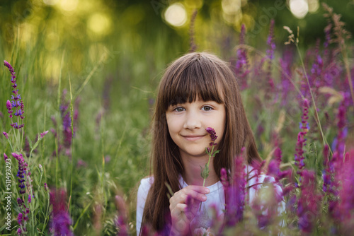 Cute girl 7-8 years, sitting in the lavender field with a flower in her hand, looking at the camera