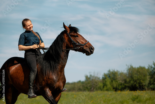 Smiling woman training young horse in field. Female rider pulling up reins trotter in rural.