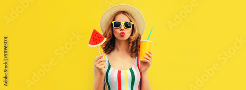 Summer colorful portrait of beautiful young woman blowing her lips with cup of juice and lollipop or ice cream shaped slice of watermelon wearing straw hat on yellow background, blank copy space