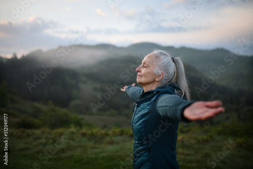 Senior woman doing breathing exercise in nature on early morning with fog and mountains in background.