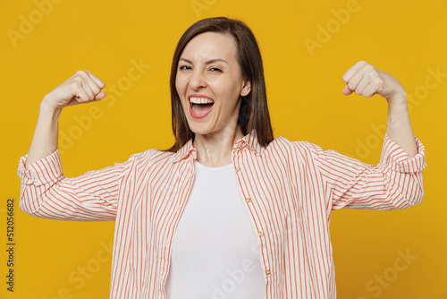 Young excited fun strong sporty fitness woman she 30s wears striped shirt white t-shirt showing biceps muscles on hand demonstrating strength power isolated on plain yellow background studio portrait