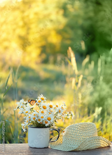 chamomile flowers in white cup and braided hat on table in garden, sunny natural abstract background. summer season. composition. relaxation, harmony atmosphere