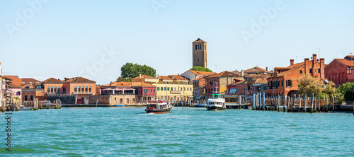 Cityscape of Murano island, famous for the production of artistic glass. Sea channel with two ferry boats, bell tower of the Cathedral, Basilica of saints Maria and Donato, VII century. Venice, Italy.