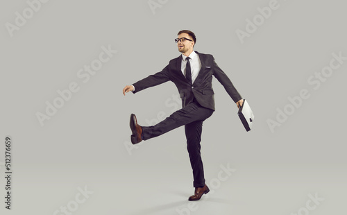 Businessman with briefcase and crazy expression ridiculously goes to work in company's office. Man in suit is making wide and big strides towards his career. Isolated on gray background. Full length.