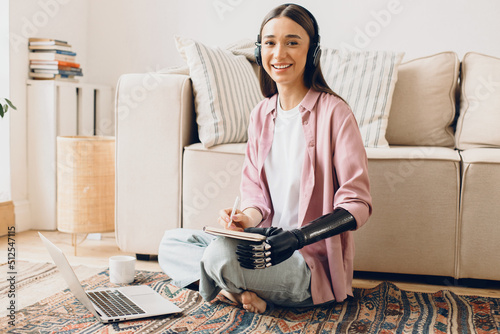 Smiling cheerful young female student listening to online English lesson in her wireless earphones, noting down in copybook, having bionic cyber hand, sitting against white sofa, in front of laptop