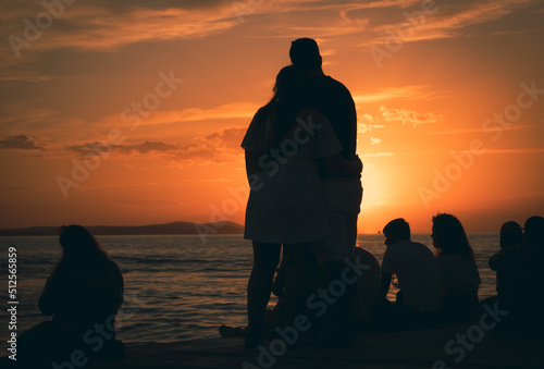 Zakochana para przytula się i patrzy na zachód słońca. Ludzie siedzący na promenadzie patrzący na zachód słońca. Zdjęcie pod światło, zarys sylwetek.