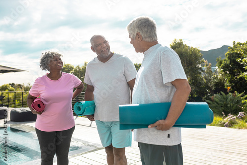 Happy multiracial senior friends with exercise mats talking while standing against sky in yard