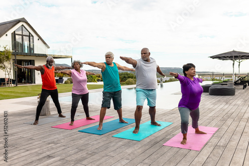 Multiracial senior friends practicing warrior 2 pose on mats at poolside against sky in yard