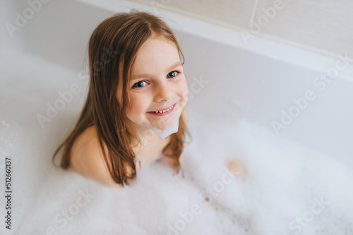 A small, smiling, beautiful red-haired girl with long hair, the child bathes, washes in a white bath with soap foam and hot water. Happy childhood.