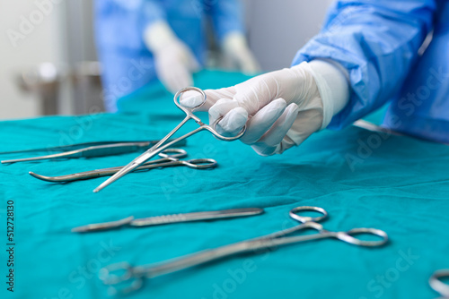 Nurse hand taking surgical instrument for group of surgeons at background operating patient in surgical theatre. Steel medical instruments ready to be used. Surgery and emergency concept