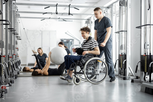People exercising at rehabilitation center, rehabilitologist walking with guy in a wheelchair. Concept of kinesiology and recovery from injuries