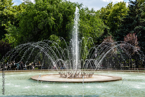 Beautiful cascading fountain against blurred background of deciduous trees. Fountain with huge bowl in shady landscaped park for relaxation and walks. City center. Krasnodar, Russia - June 16, 2022