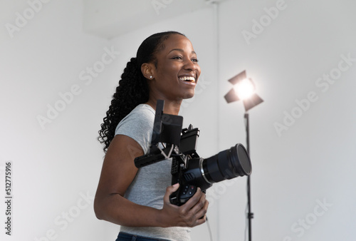 African American Female Videographer Posing with Video Camera in Hand on Film Set. Camera Woman is Proud and Smiling Looking Off Camera.