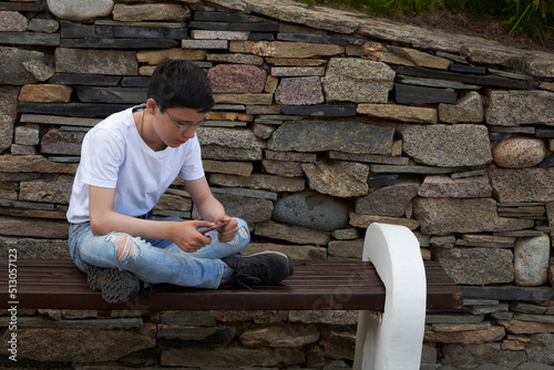 A dark-haired teenage boy with glasses is sitting on a bench and playing on a smartphone.