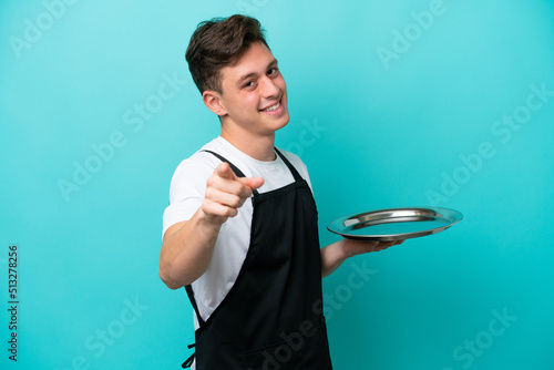 Young waitress with tray isolated on blue background pointing front with happy expression