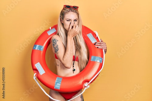Young caucasian woman wearing bikini and holding lifeguard float smelling something stinky and disgusting, intolerable smell, holding breath with fingers on nose. bad smell