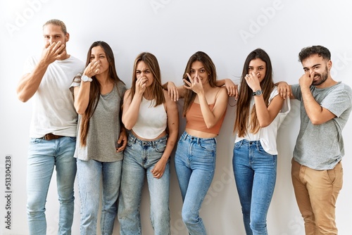 Group of young friends standing together over isolated background smelling something stinky and disgusting, intolerable smell, holding breath with fingers on nose. bad smell