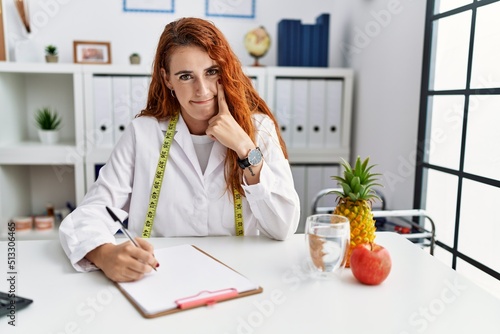 Young redhead woman nutritionist doctor at the clinic pointing to the eye watching you gesture, suspicious expression